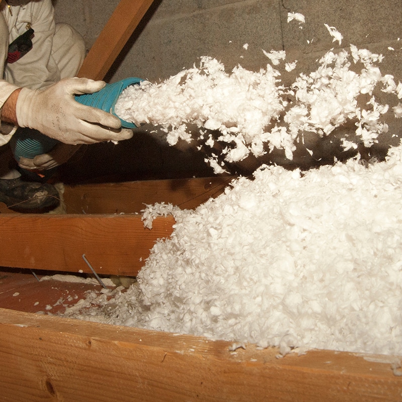 Un ouvrier, ganté de blanc, souffle de l'isolant en cellulose soufflé blanc dans un grenier entre des poutres en bois.