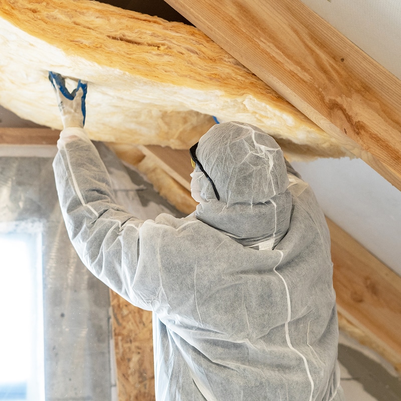 Ouvrier avec combinaison blanche, lunettes et gants installant de la laine de verre isolante entre des chevrons de bois dans un grenier.