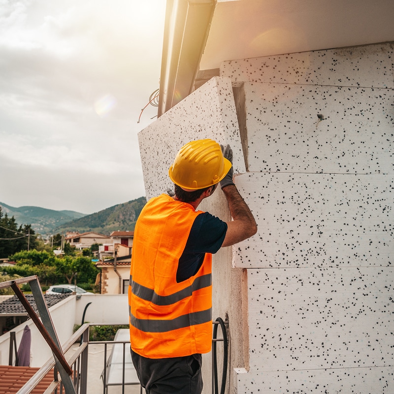 Ouvrier portant un casque jaune et un gilet orange installant des panneaux isolants blancs (polystyrène) sur un mur extérieur, avec un paysage vallonné en arrière-plan.