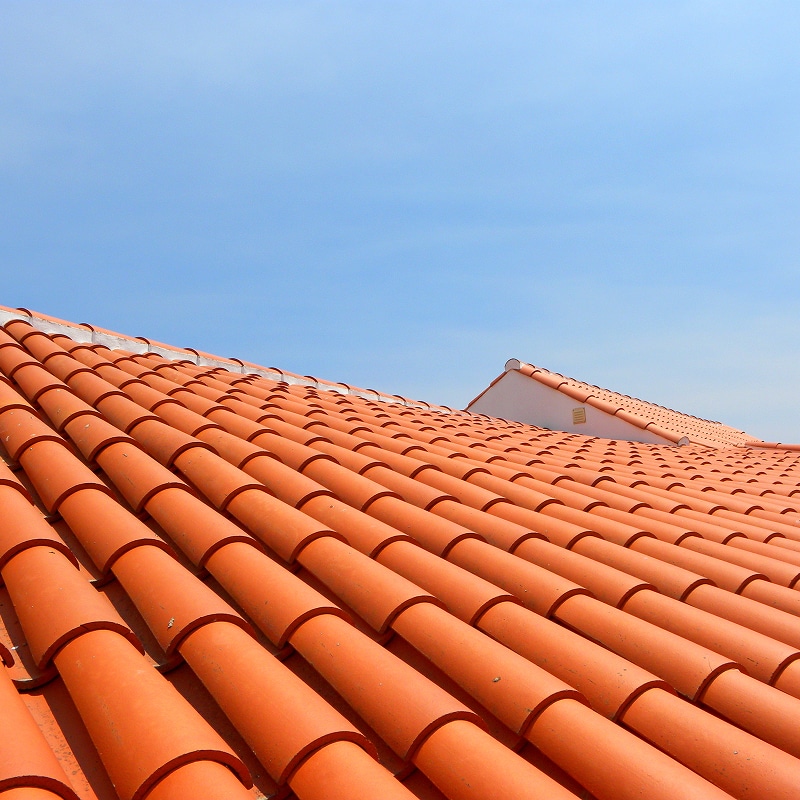 Tuiles en terre cuite, couleur orange vif, formant un toit en pente sous un ciel bleu clair, avec une lucarne blanche visible au loin.